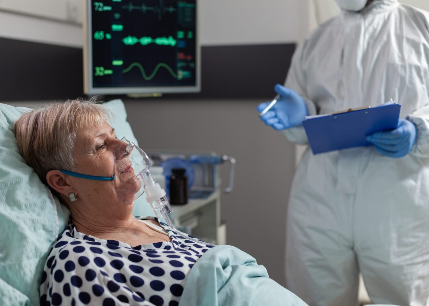 Sick senior woman inhale and exhale through oxygen mask laying in hospital bed during global pandemic with coronavirus. Doctor dressed in coverall pee suit to prevent infection with covid-19.